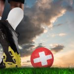 Composite image of football boot kicking swiss ball against green grass under blue and orange sky